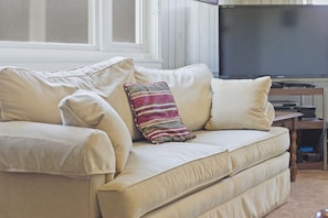 A beige sofa with multiple cushions, a television on a stand, and a window with blinds.