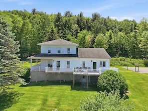 A two-story house with a covered porch and a small deck, surrounded by lush greenery.