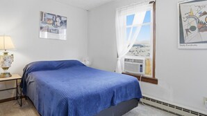 Une chambre à coucher avec un lit, une table de chevet avec une lampe, une fenêtre avec des rideaux et des œuvres d’art murales.