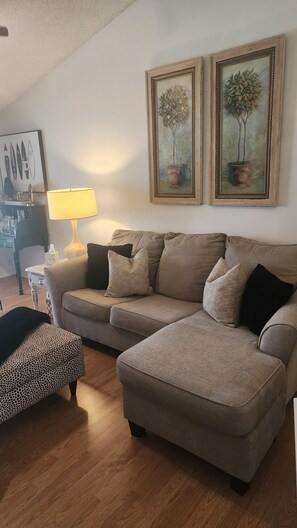 Ein Wohnzimmer mit einer Couch, einem gemusterten Hocker, einer Lampe und zwei gerahmten Bildern an der Wand.