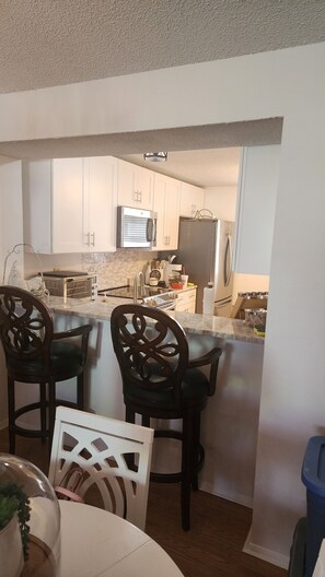 A modern kitchen with white cabinets, a built-in microwave, and a stainless steel refrigerator. There are two wooden chairs at the island and a white table with a glass dome centerpiece. - Florida Fun Villa in the heart of Oldsmar between Tampa and Clearwater (Oldsmar)