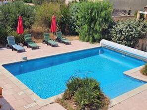 A rectangular swimming pool with a tiled edge, surrounded by a paved patio with lounge chairs and red umbrellas. - Waterfront house with 2 bedrooms, living room, air conditioning, pool, and secure parking (Le Barcarès)