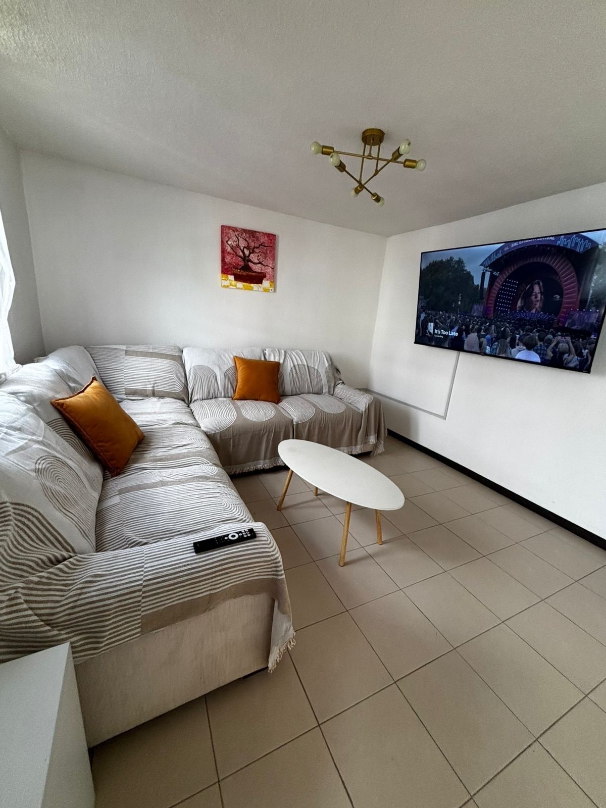 Un salon moderne avec un canapé d’angle, une petite table blanche, un téléviseur à écran plat fixé au mur et un luminaire suspendu au plafond.
