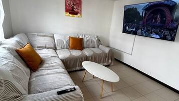 Un salon moderne avec un canapé d’angle, une petite table blanche, un téléviseur à écran plat fixé au mur et un luminaire suspendu au plafond.