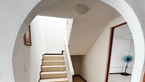Uma escada com corrimão branco que leva a um patamar, um ventilador azul no chão e uma porta no final do corredor.