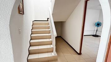 Un escalier avec une rampe blanche menant à un palier, un ventilateur bleu posé sur le sol et une porte au bout du couloir.