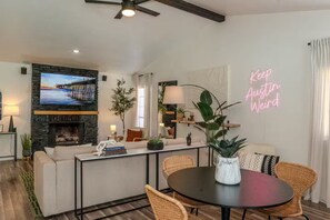 A modern living room with a fireplace, a television mounted on a stone wall, a dining area with a round table, and a neon sign on the wall. - Austin Home with Private Pool & Spa (Austin)
