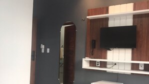 Quarto de hotel moderno com uma cama, uma TV de tela plana presa a uma parede de madeira, um frigorífico branco pequeno e um espelho.