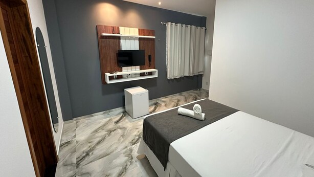 Quarto de hotel moderno com uma cama, um pequeno frigorífico branco, uma televisão fixada num painel de madeira e uma casa de banho com cortina.