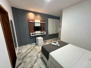Een moderne hotelkamer met een bed, een kleine witte koelkast, een televisie bevestigd aan een houten paneel, en een badkamer met een gordijn.