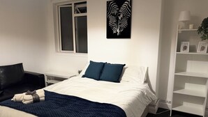 A bedroom with a bed, a window, a wall-mounted artwork, a bedside table, a lamp, and a shelf with decorative items. - Modern City Centre Apartment | Free Parking | Self Check-In (Southampton)