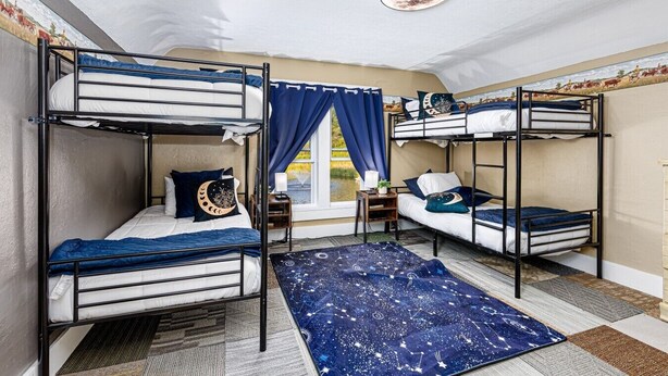 Ein Schlafsaalzimmer mit Etagenbetten, einem kleinen Tisch, einem Nachttisch, einem Fenster mit Vorhängen und einem Teppich mit Sternenmuster.