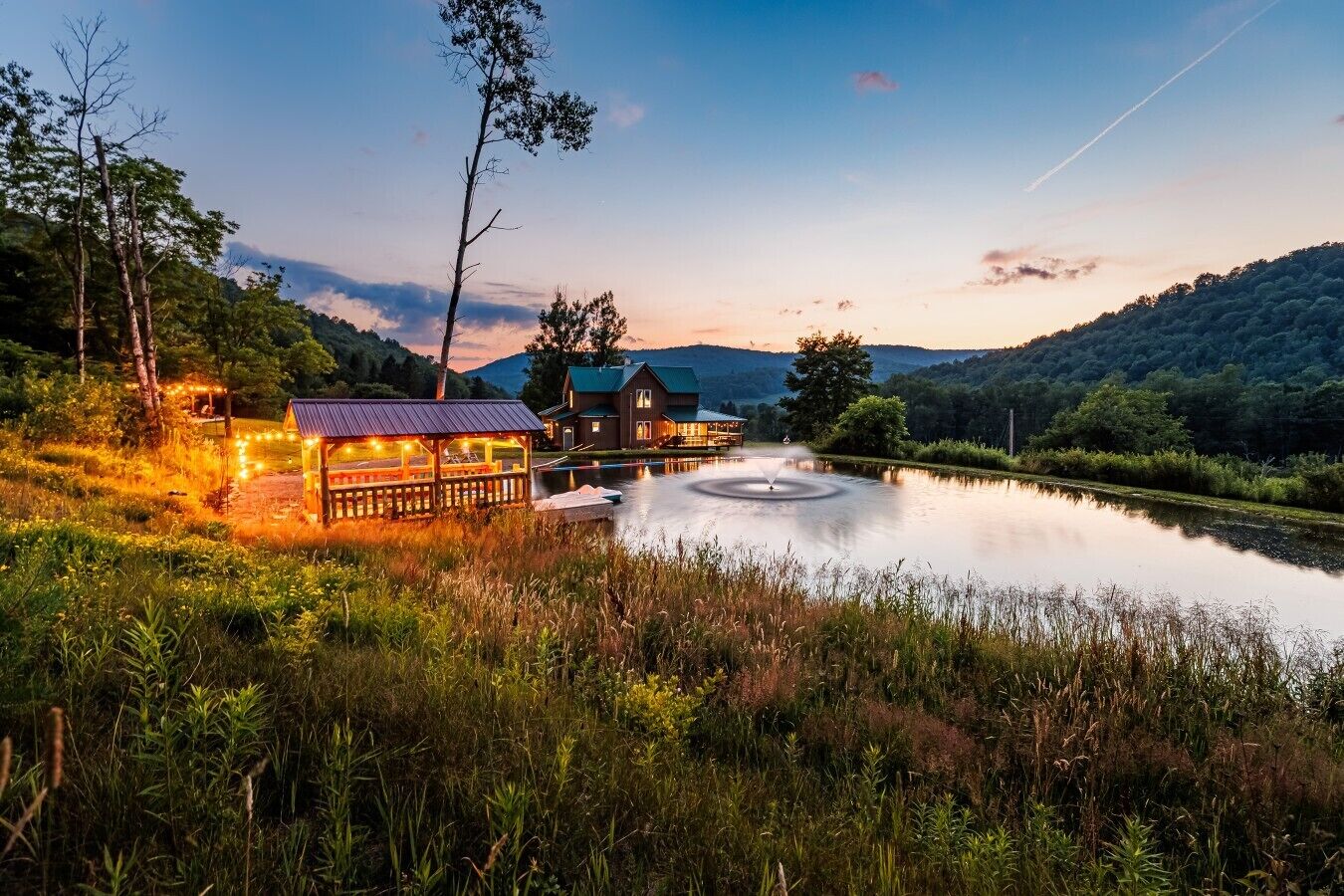 A lakeside lodge with a wooden deck, a fountain, and a house with a green roof.
