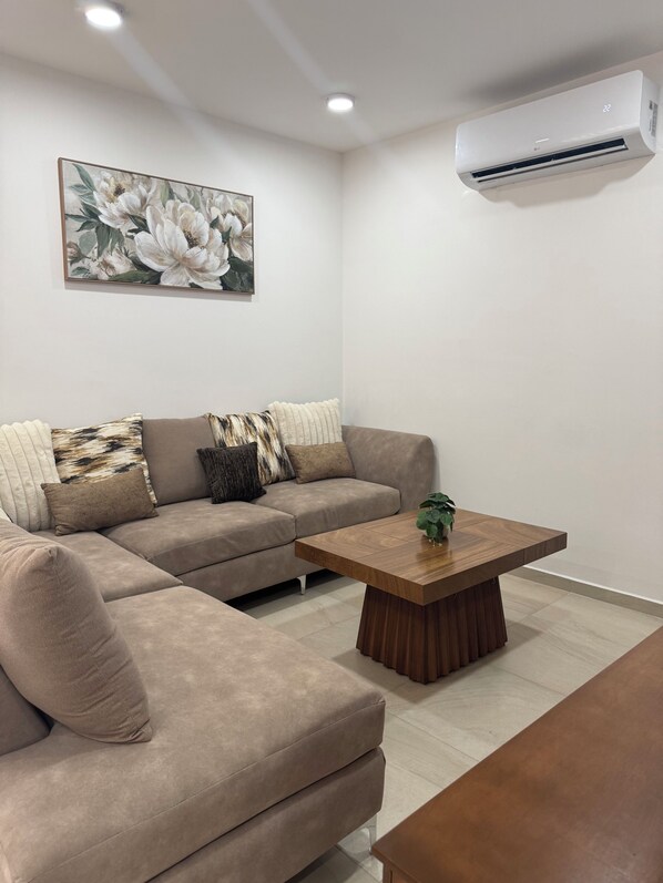 A modern living room with a beige sectional sofa, a wooden coffee table, and a wall-mounted air conditioner.