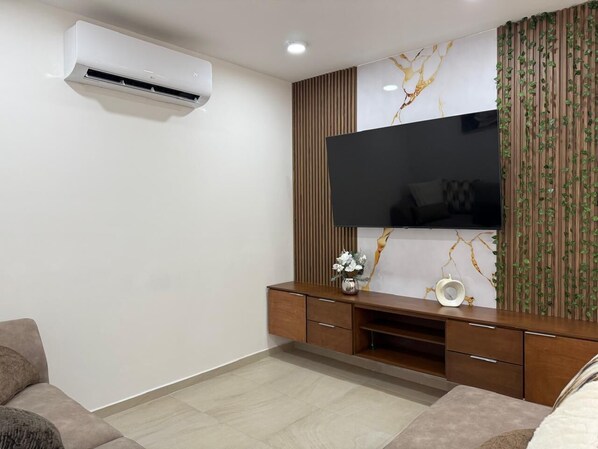 A modern living room with a flat-screen TV mounted on a wall with a marble-like design, a wooden entertainment unit below, and an air conditioning unit on the wall.