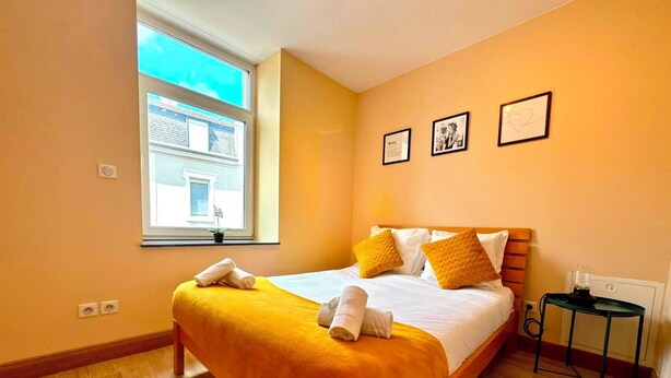 A hotel room with a large bed, a window with a view of buildings, a small table with a lamp, and a wall with framed pictures. - Spacious 1 bedroom with 4 beds  (Luxembourg)