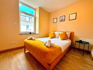 A hotel room with a large bed, a window with a view of buildings, a small table with a lamp, and a wall with framed pictures. - Spacious 1 bedroom with 4 beds  (Luxembourg)