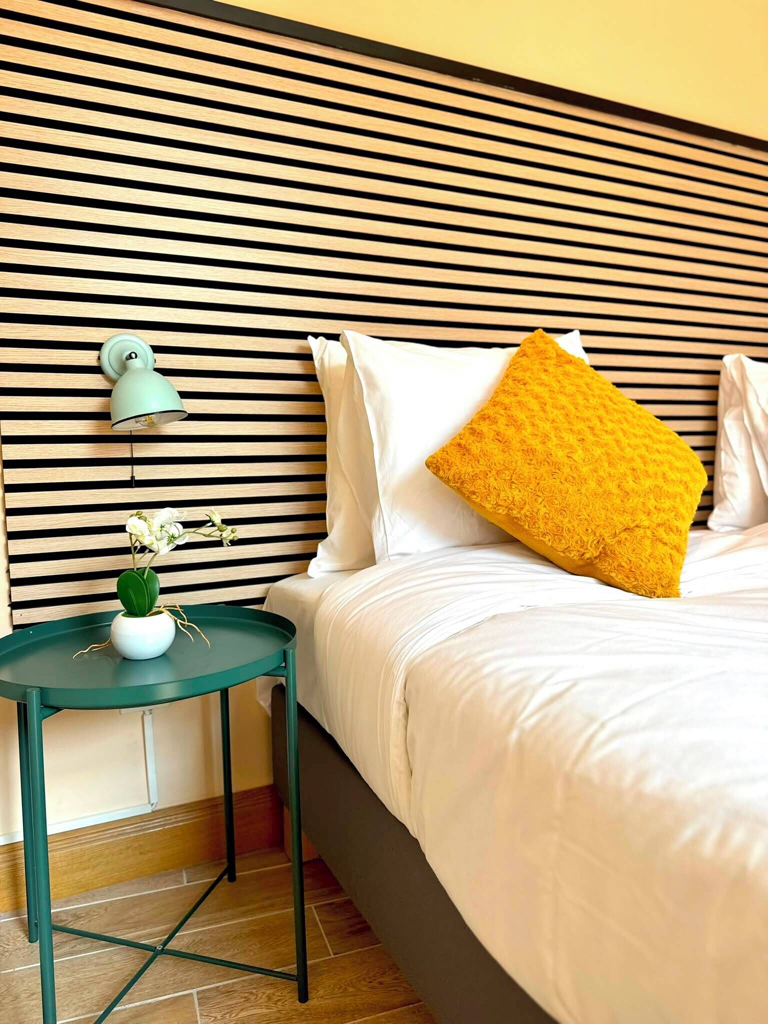 Een keurig opgemaakt bed met witte lakens en één geel kussen, een klein groen tafeltje met een witte vaas, en een wand met horizontale houten latten.
