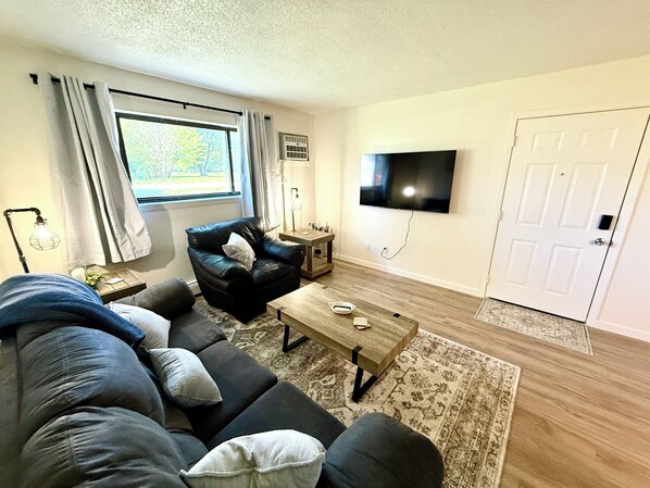 A living room with a sofa, a coffee table, a TV mounted on the wall, and a door.