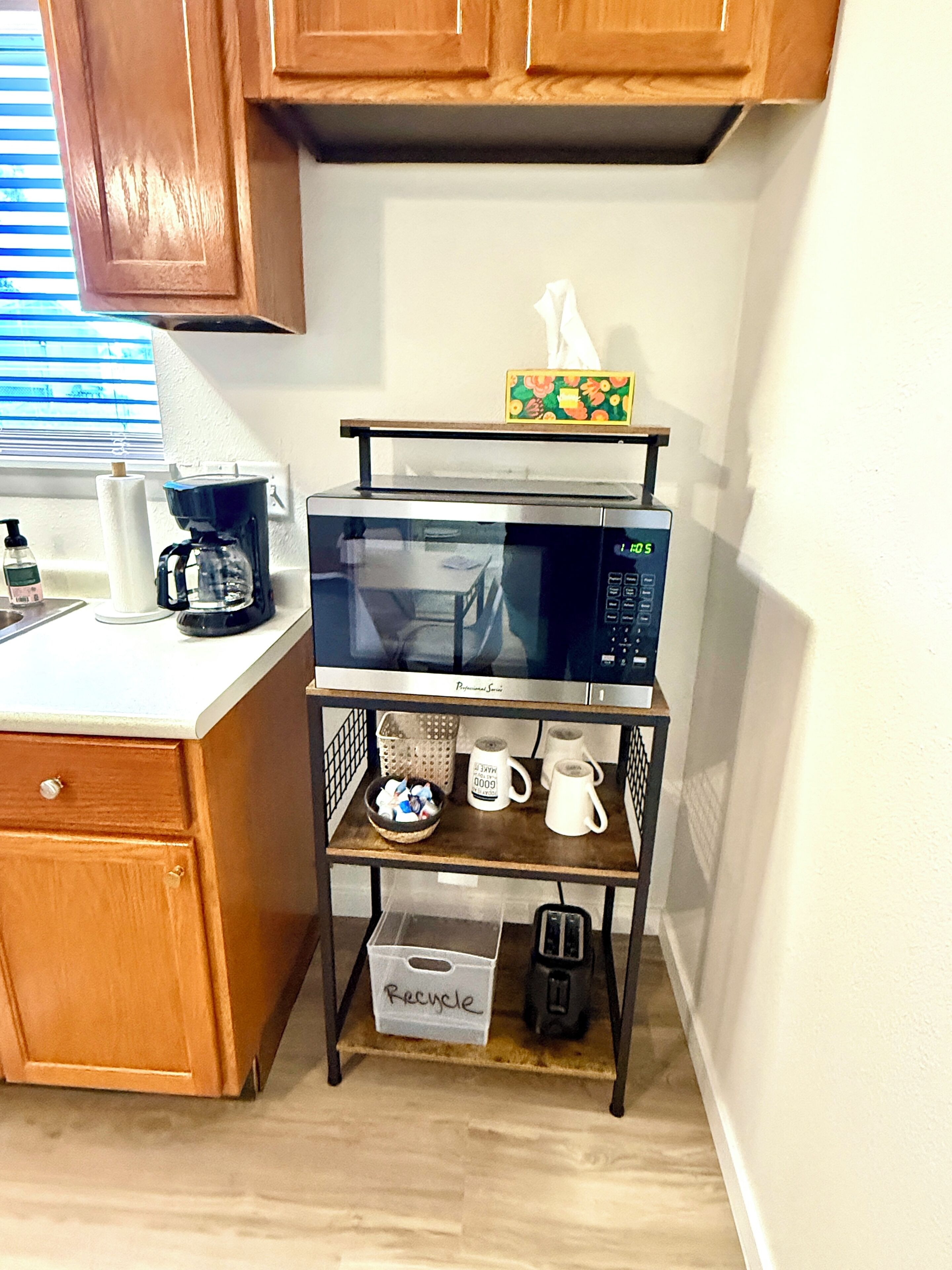 A kitchen with wooden cabinets, a microwave on a metal shelf, a coffee maker, and a basket labeled 'Recycle'.