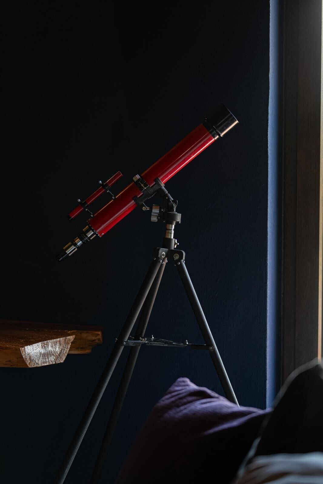 A telescope on a tripod in a room with a dark wall and a window.