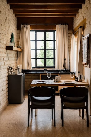 A dining area with a brick wall, wooden beams, a window with curtains, a table set for two, and a refrigerator.