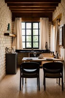 Un comedor con pared de ladrillo, vigas de madera, una ventana con cortinas, una mesa preparada para dos personas y un refrigerador.