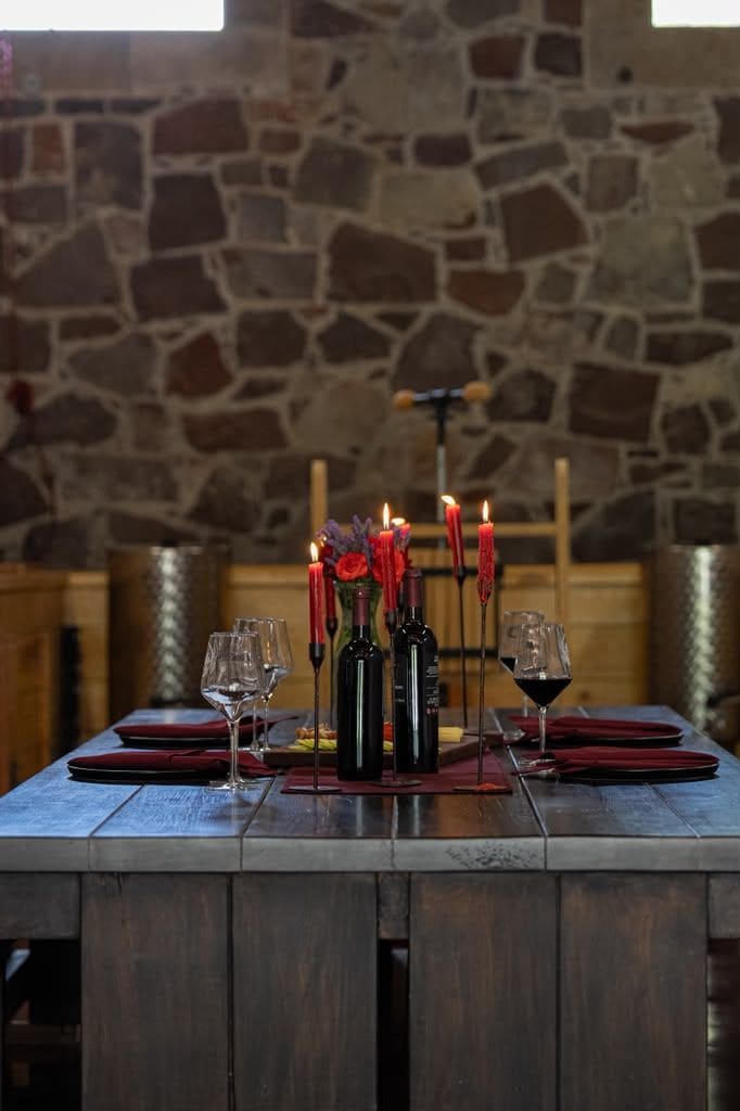 A dining table set with wine glasses, bottles, and candles in a room with a stone wall.