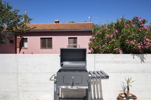 Un barbecue est installé devant un bâtiment rose, doté d’un toit en tuiles et d’un mur blanc.