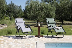 Zwei Liegestühle mit einem Tisch und einer Weinflasche dazwischen, auf einer Steinterrasse mit einem Pool im Vordergrund.