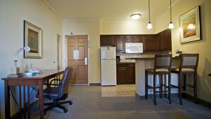 Une cuisine compacte équipée d’un réfrigérateur, d’un four à micro-ondes et de meubles de rangement en bois. On y trouve un coin repas avec une table et des chaises, ainsi qu’un petit espace bureau avec un bureau et une chaise.