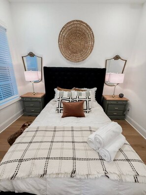 A neatly made bed with a checkered bedspread, two nightstands with lamps, a round woven wall hanging, and a mirror on each nightstand. - Luxury Designer Home | Steps to NoDa | City Views (Charlotte)