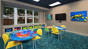 Ein Kinderspielbereich mit bunten Stühlen und einem Tisch, großen Fenstern und Wandbildern.