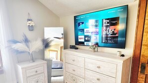 A living room with a flat-screen TV mounted on the wall, a wooden dresser with drawers, a small potted plant, and decorative items.