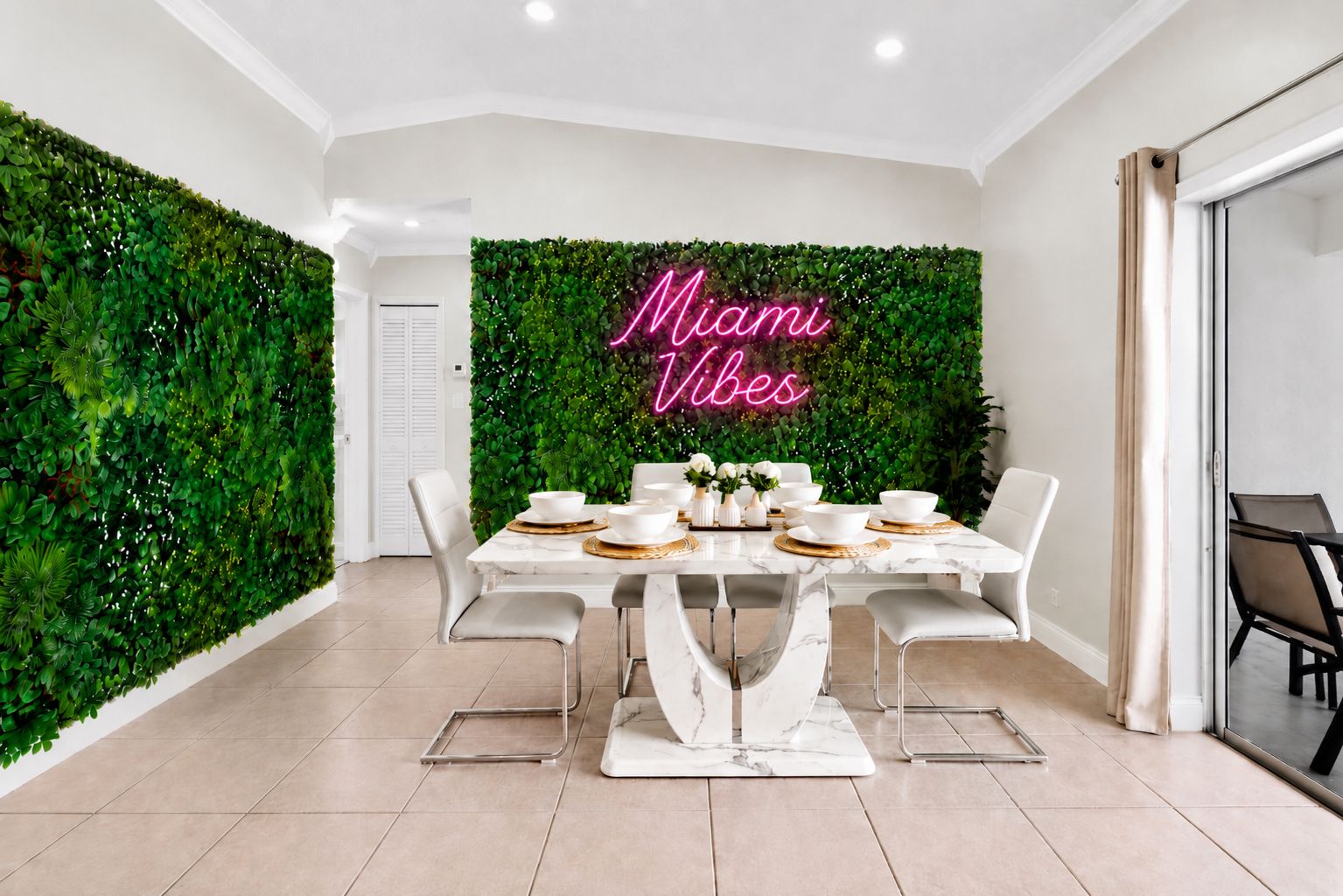 A dining area with a table set for four, surrounded by white chairs, against a backdrop of a green wall with a neon sign.