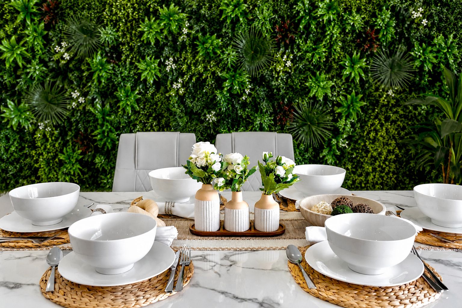 A table set with white bowls and plates, accompanied by woven placemats and silverware, against a backdrop of lush green foliage.