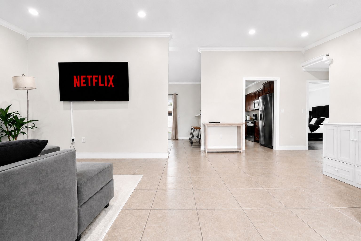 A modern living room with a flat-screen TV mounted on the wall, a grey sofa, and a kitchen area in the background.