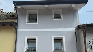 Un edificio di due piani con facciata grigia, modanature bianche e numerose finestre. Sono presenti due condizionatori e una pianta in vaso al piano terra.