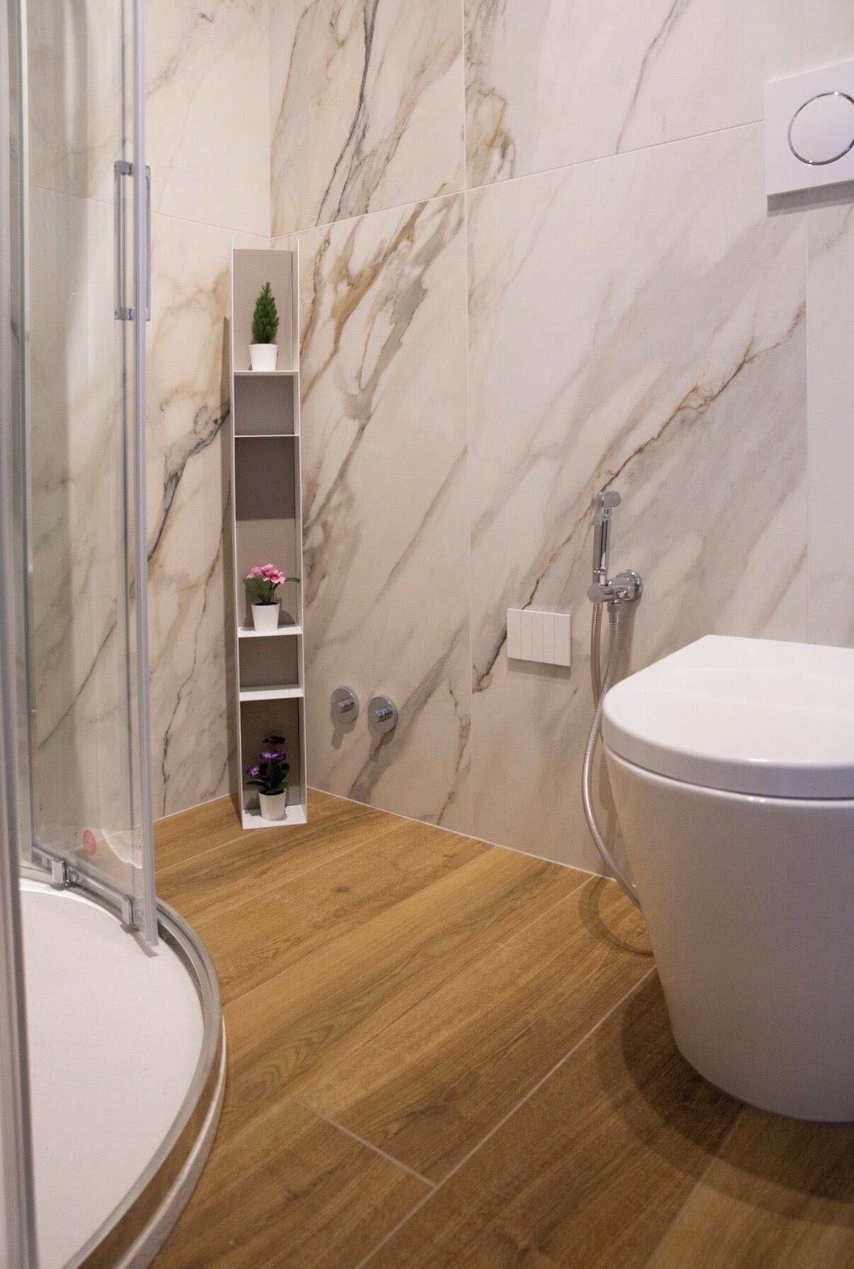 A bathroom with a marble wall, a wooden floor, a toilet, a shower, and a shelf with plants.
