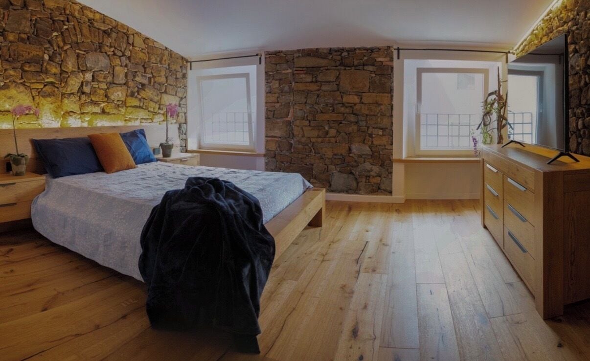 Ein Schlafzimmer mit Steinwand, einem Bett mit Kissen, einer hölzernen Kommode und einem Fenster.