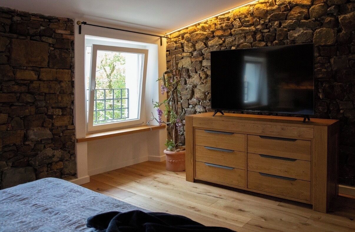 A room with a stone wall, a large flat-screen TV, a wooden cabinet, a window with a view, and a bed.