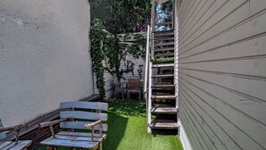 Un espacio exterior reducido con un banco de madera, una pequeña mesa, una zona de césped y una escalera metálica que sube a un edificio.
