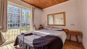 Un dormitorio con una cama grande, un espejo, una mesita de noche y una ventana con vistas a edificios.