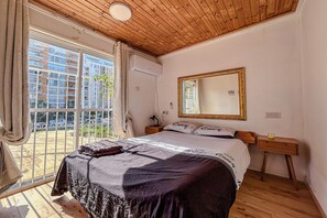 A bedroom with a large bed, a mirror, a bedside table, and a window with a view of buildings. - Bright 2BR Home with Private Garden and Free Parking (Herzliya)
