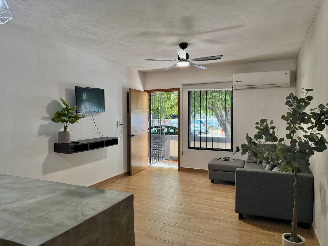 A modern living room with a flat-screen TV, a ceiling fan, a grey sofa, and a wooden floor.
