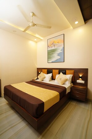 A hotel room with a large bed, a ceiling fan, a painting on the wall, and a nightstand with a lamp. - Hotel Royace Executive By Glitz Hotels (Mumbai)