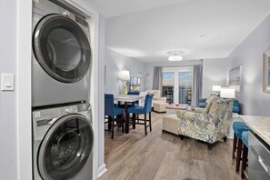 Ein modernes Apartment mit integrierter Waschmaschine und Trockner, einem Essbereich mit blauen Stühlen, einem Sofa mit floralem Muster und einem großen Fenster mit Blick auf die Stadt.