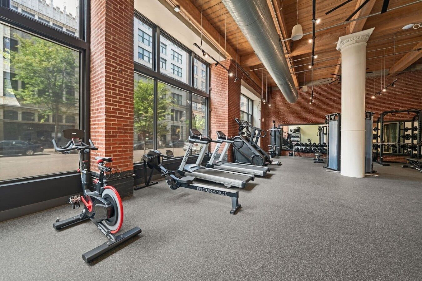 Un gimnasio con equipo de ejercicio, ventanas amplias y una pared de ladrillo.