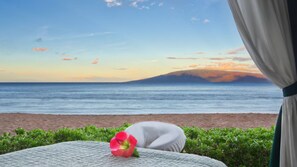 Un lit avec un oreiller blanc et une fleur rose, donnant sur une plage avec vue sur la mer et une montagne lointaine, au moment du coucher du soleil.