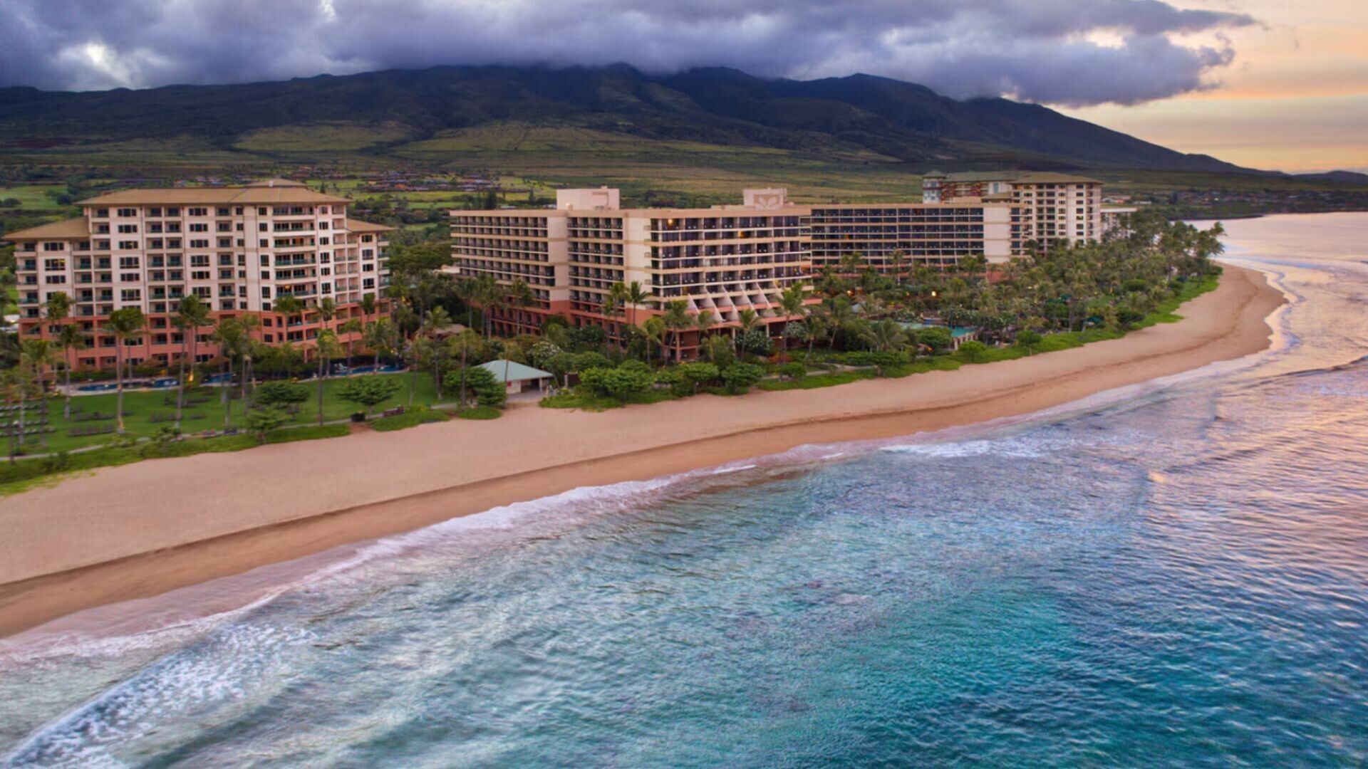Ein Strandhotel mit mehreren Gebäuden, einem Sandstrand und Blick auf den Ozean sowie das dahinterliegende Gebirge.